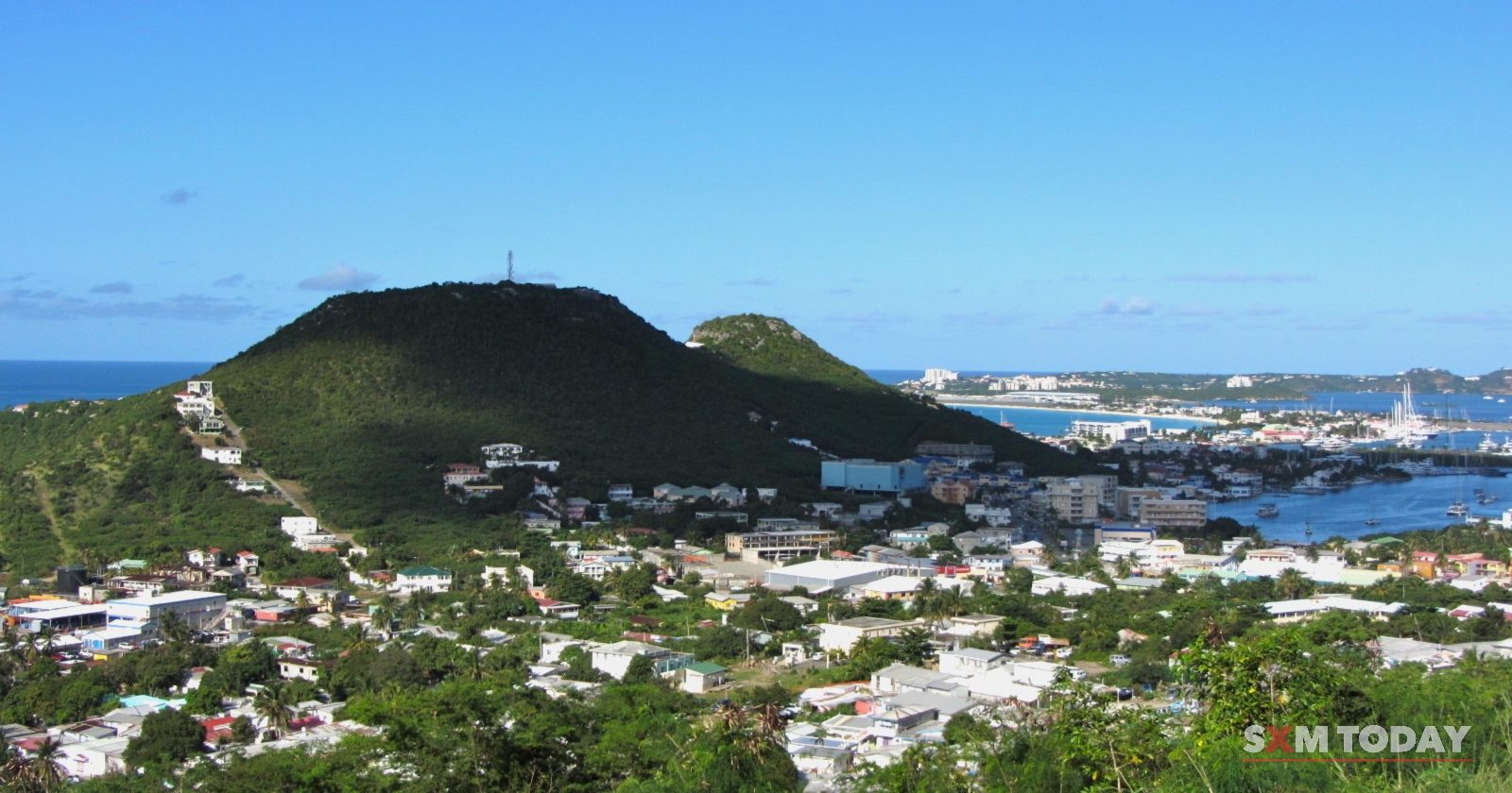 Rooftops and houses spread over green slopes below a rounded hill, with a lagoon and the sea beyond.