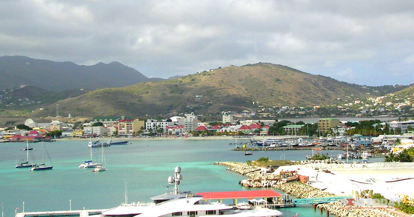 A town stretches along a bay beneath green hills, with sailboats at anchor, a rock breakwater and a red-roofed water-taxi stop in the foreground.