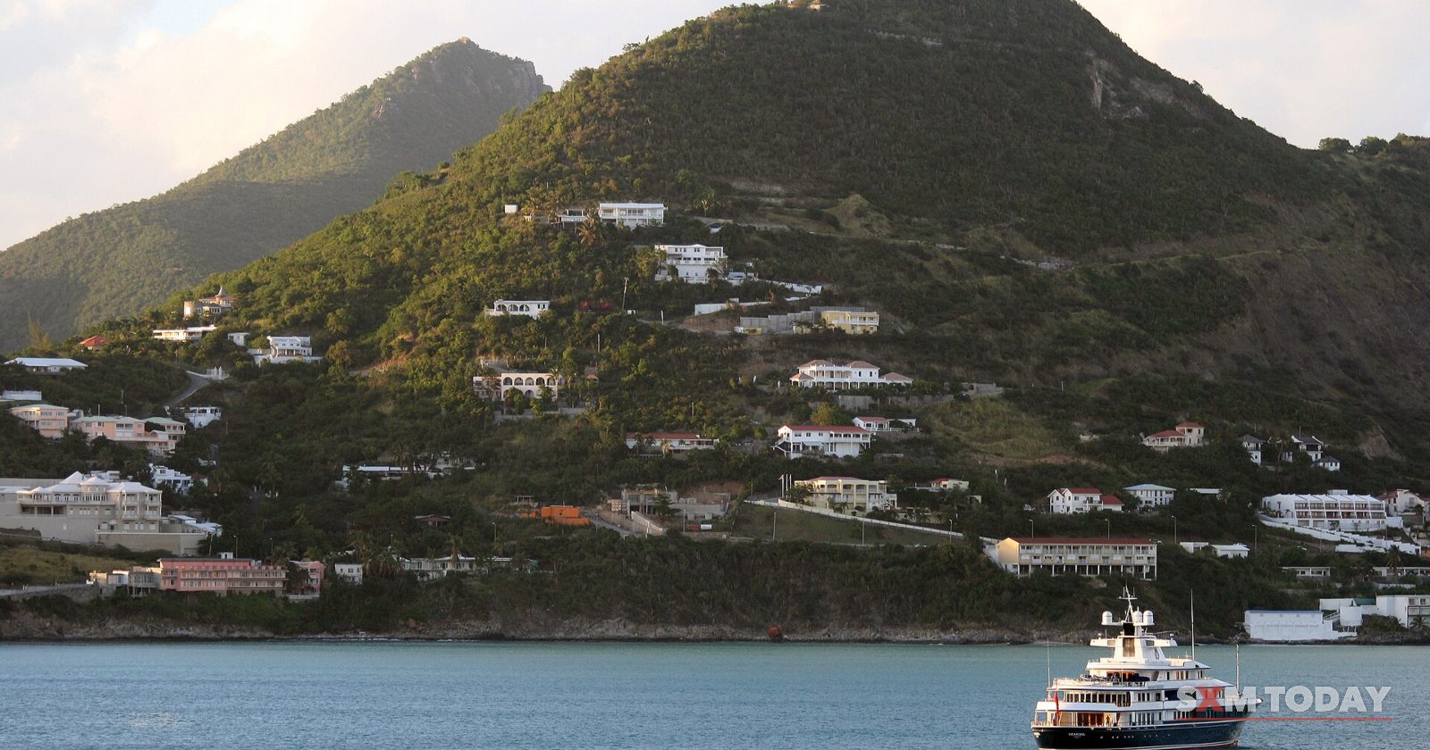 White houses dot a steep green hillside above the water, with a motor yacht moored below.