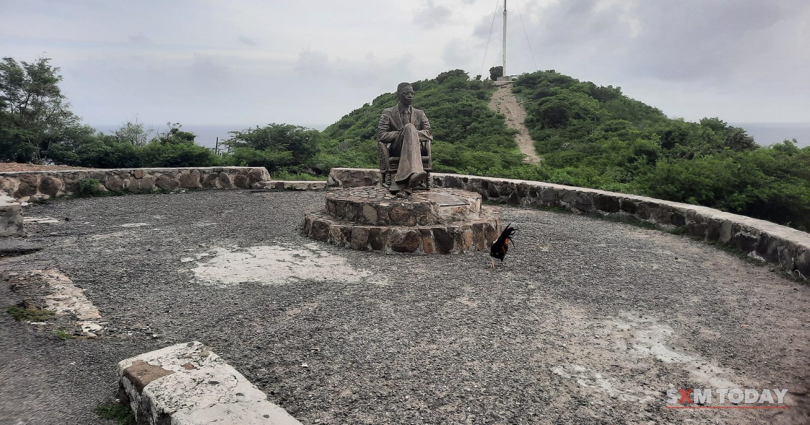 A hilltop lookout with a seated bronze statue on a stone plinth, a rooster nearby and a path up the hill behind, under grey cloud.