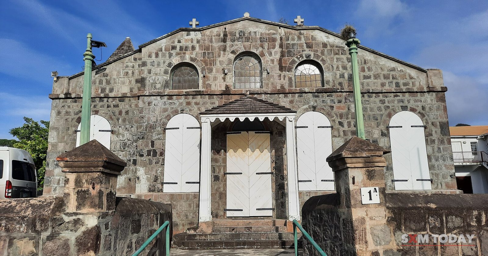 The stone front of a chapel with white shuttered doors and windows, reached by steps between stone gateposts.