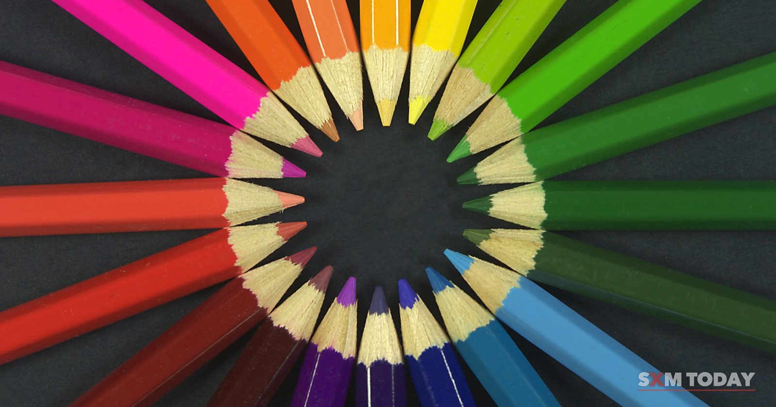 Coloured pencils arranged in a ring with their sharpened tips pointing inward, running through the colours of the rainbow on a dark background.