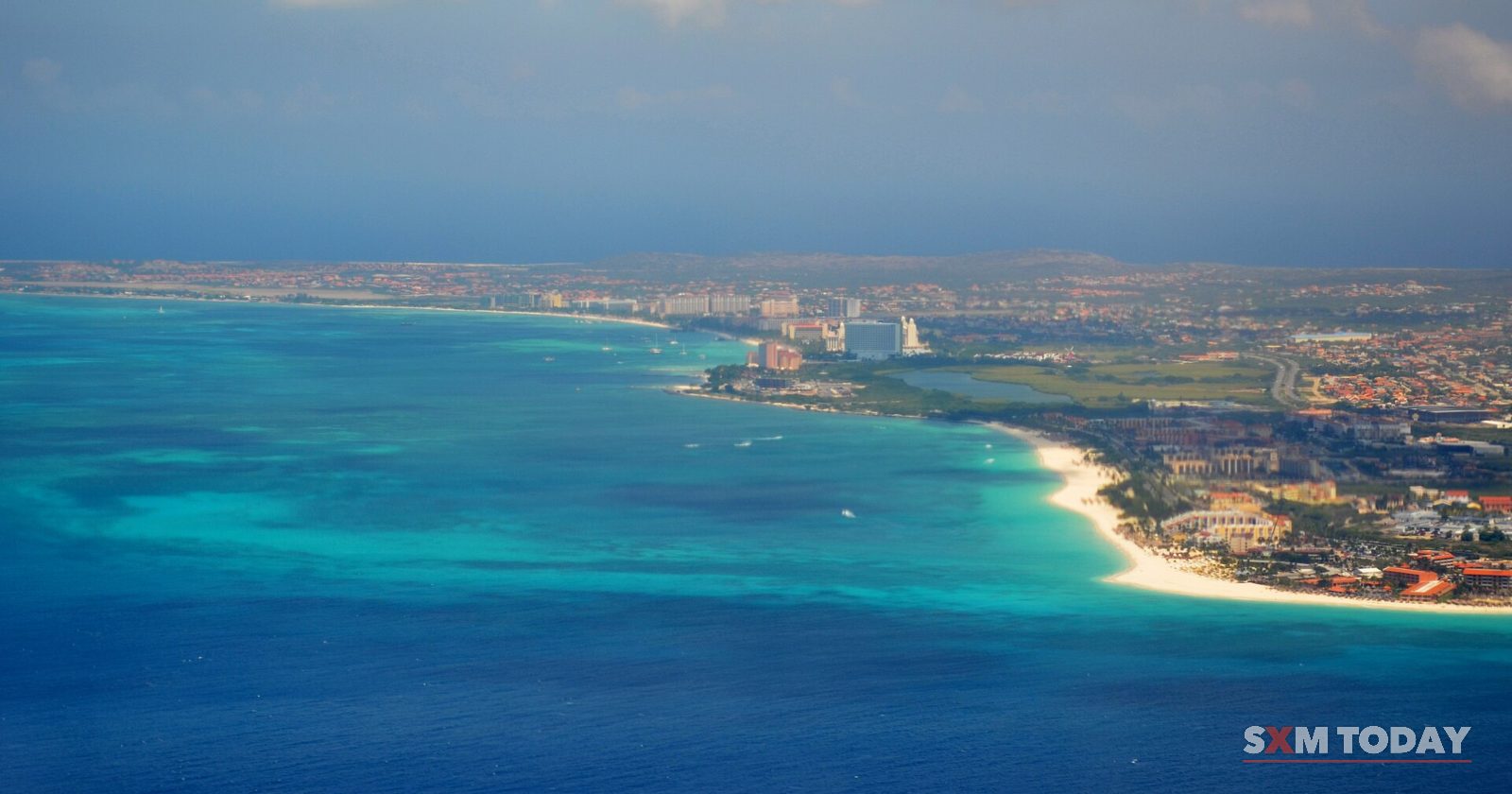 An aerial view of turquoise sea along a long white-sand coastline with high-rise hotels and a town beyond.