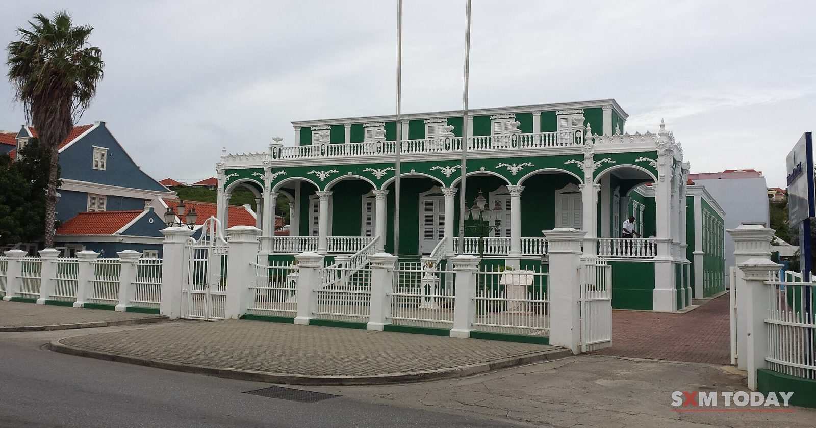An ornate green-and-white colonial building stands behind a white fence, with flagpoles in front, under a grey sky.
