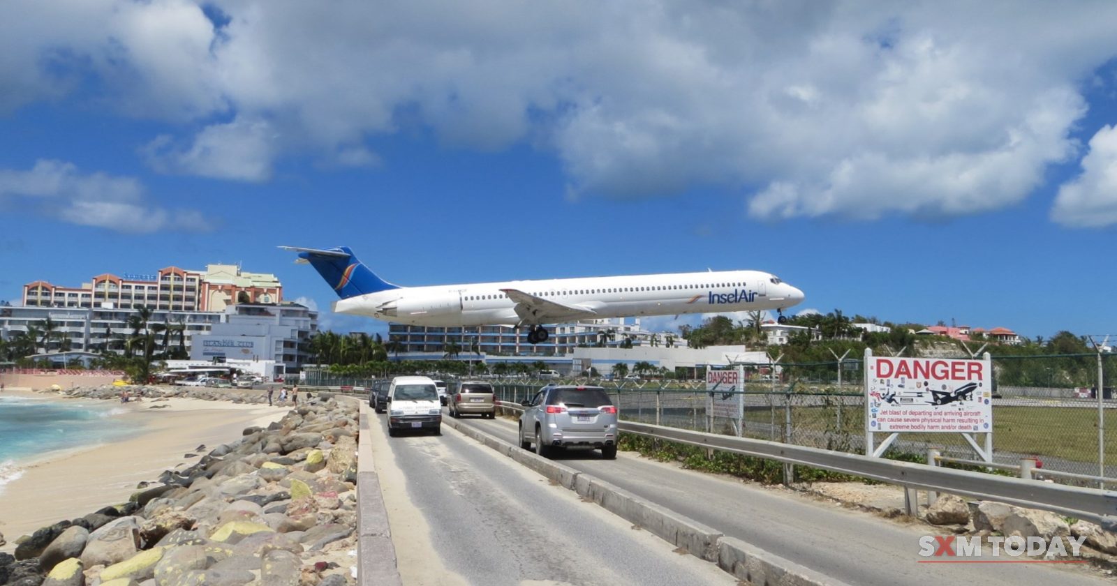 A jet flies low over a coastal road with cars and a danger sign, beside a beach and hotel buildings.