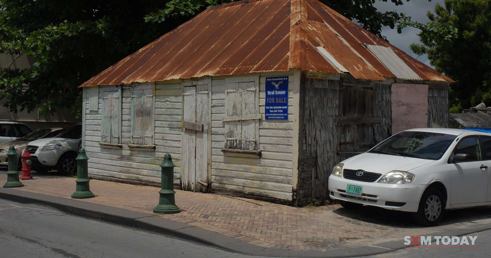 A weathered wooden house with a rusted corrugated roof stands on a street corner beside a parked white car.