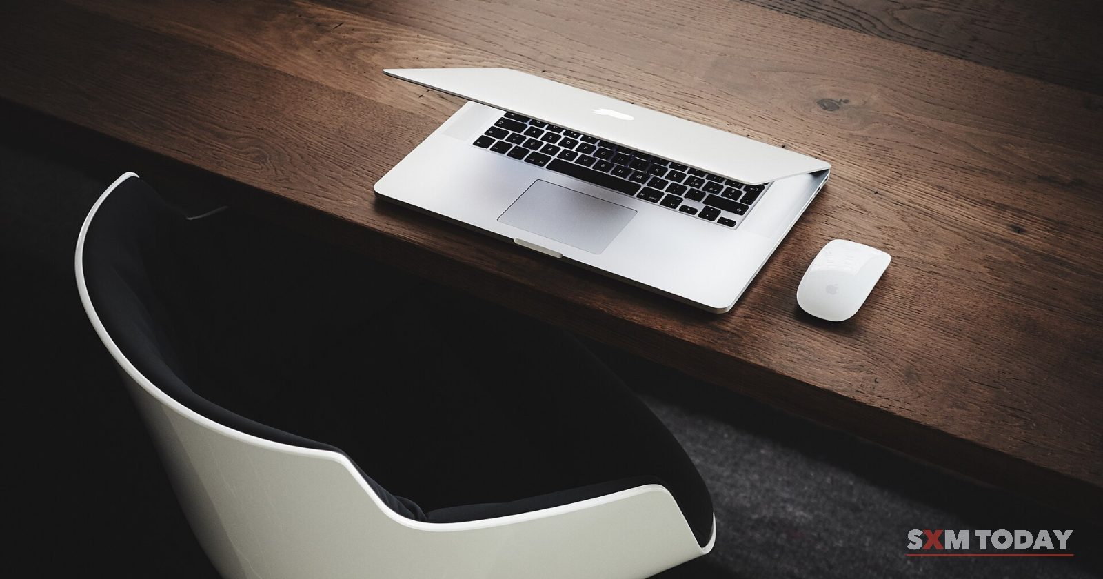 An open laptop and a mouse on a dark wooden desk beside a white chair.