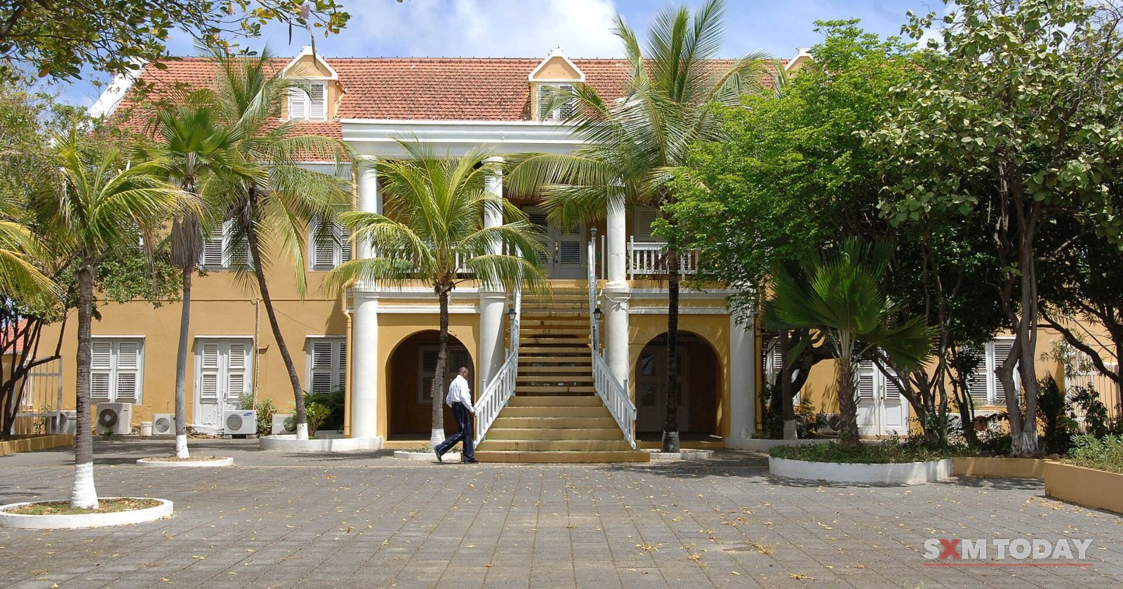 A two-storey yellow colonial government building with a white columned entrance and double staircase behind a paved square and palm trees.