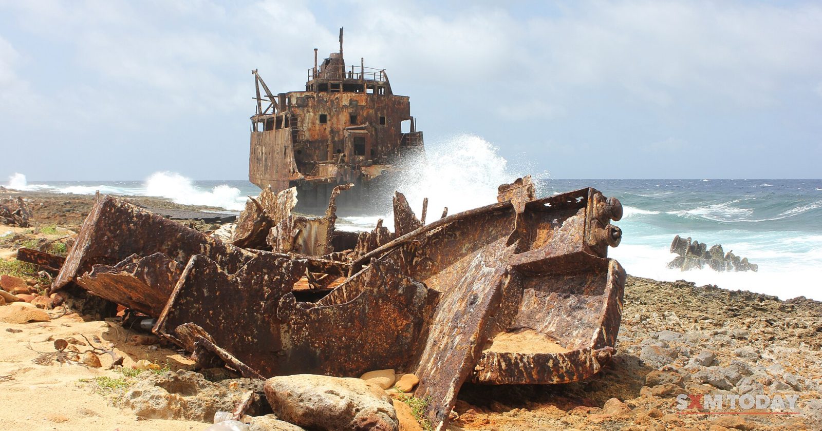 The rusted remains of a wrecked tanker lie on a rocky shore as waves break behind it.