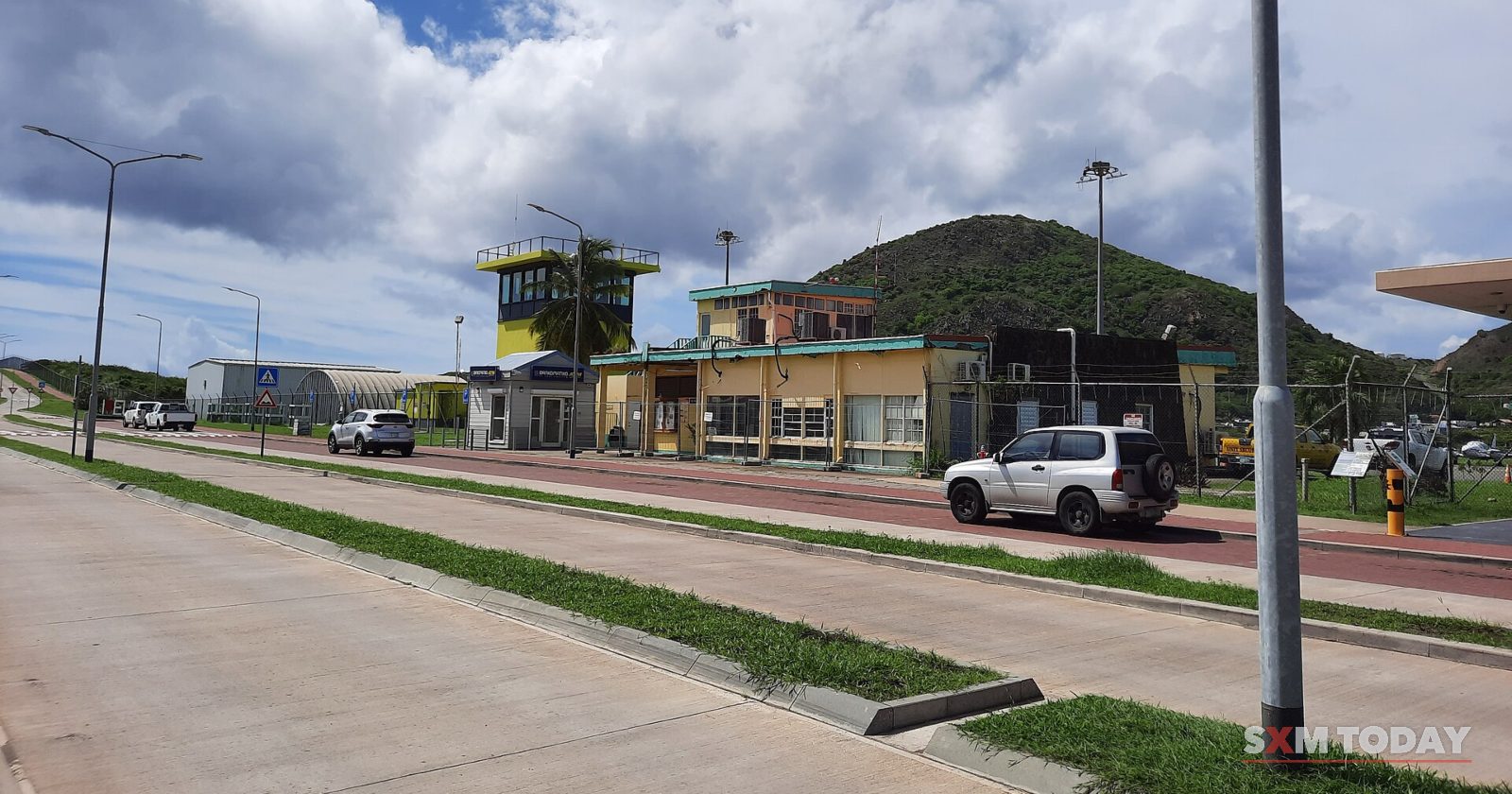 Airport buildings and a control tower line a wide road, with green hills and clouds behind.