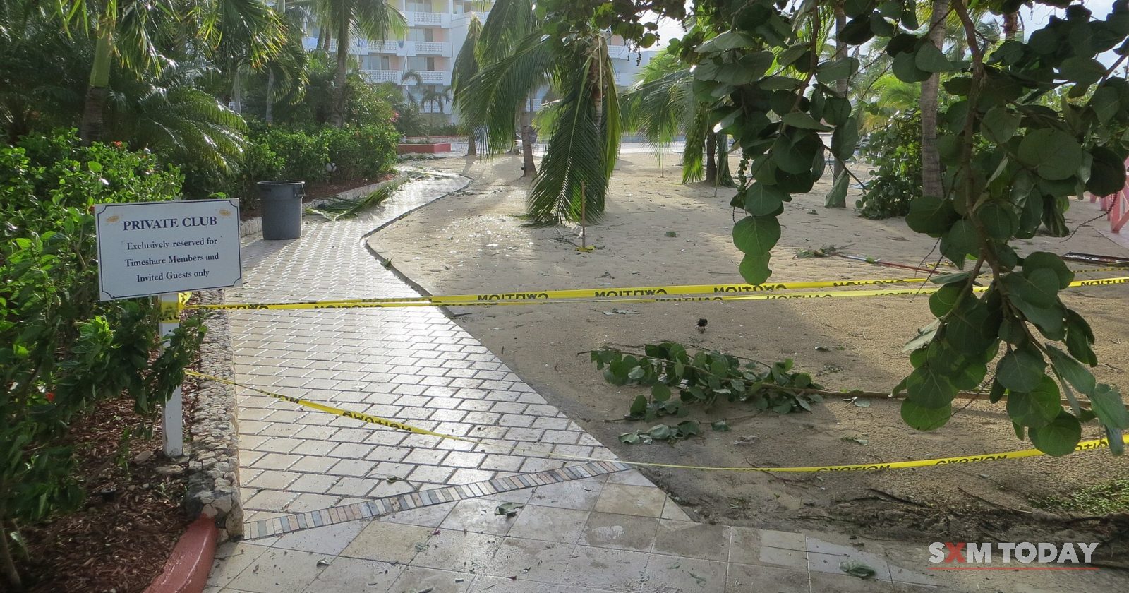 A paved path strewn with fallen branches is cordoned off with yellow tape beneath trees and a resort building.