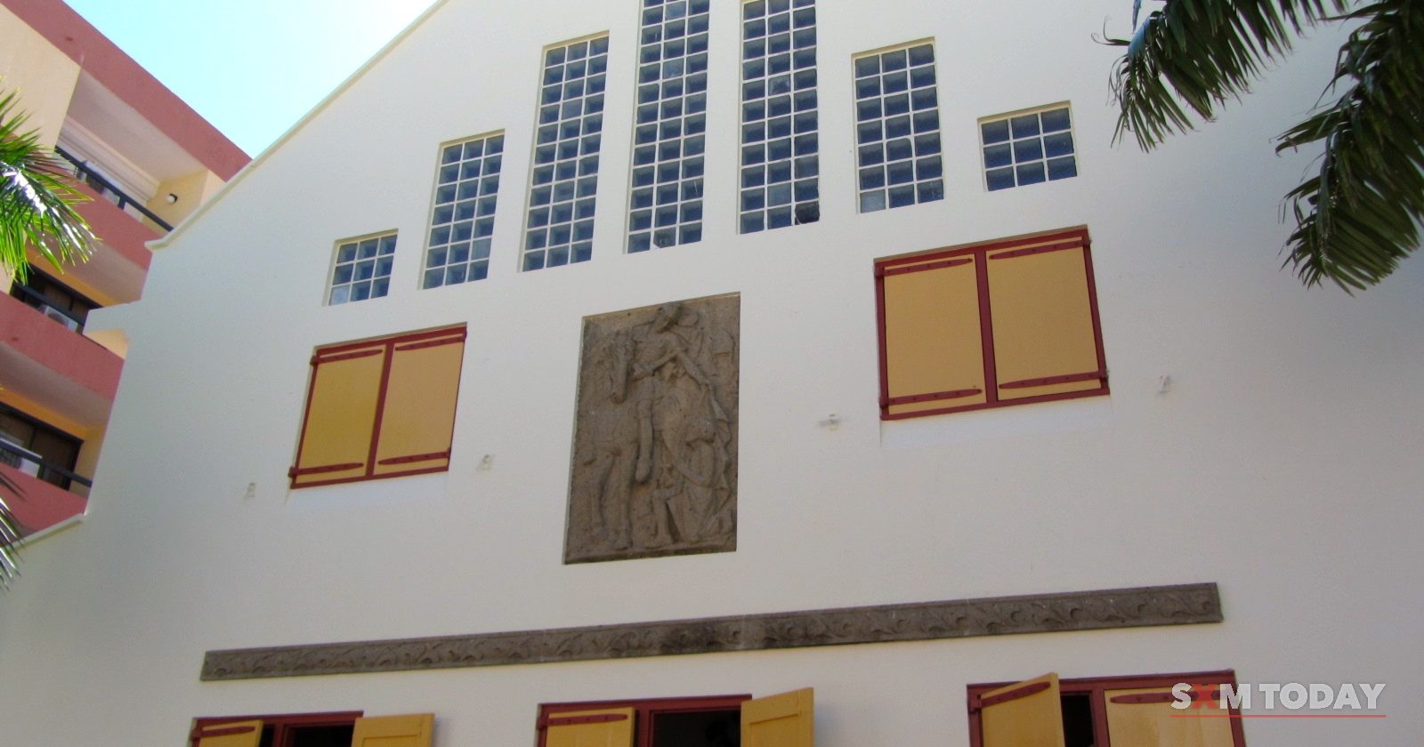 The white rear wall of a church with tall narrow windows, a carved relief panel and yellow wooden shutters, seen from below.