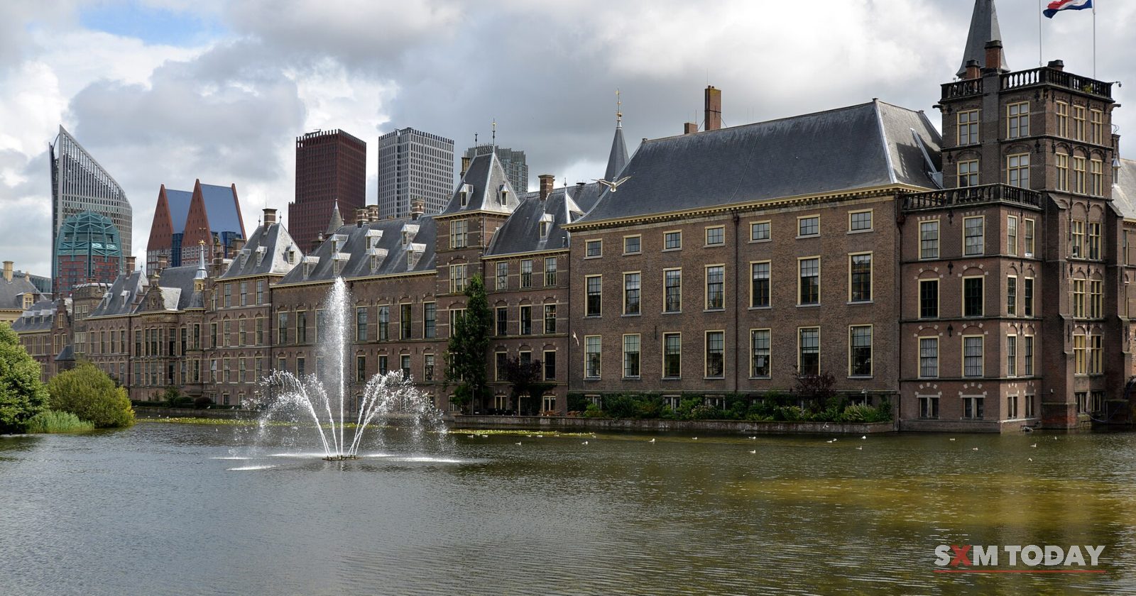 A row of brick buildings with spires and a flag across a pond with a fountain, modern towers behind under a cloudy sky.