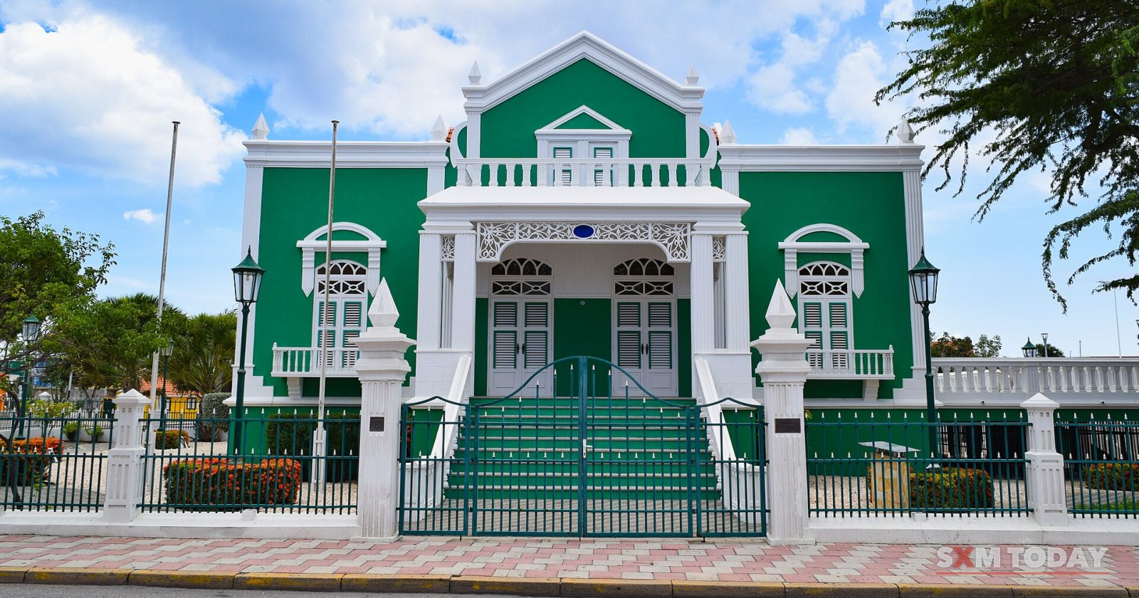A green and white colonial-style building with a central staircase and ornate gables behind a white fence, under a blue sky with clouds.