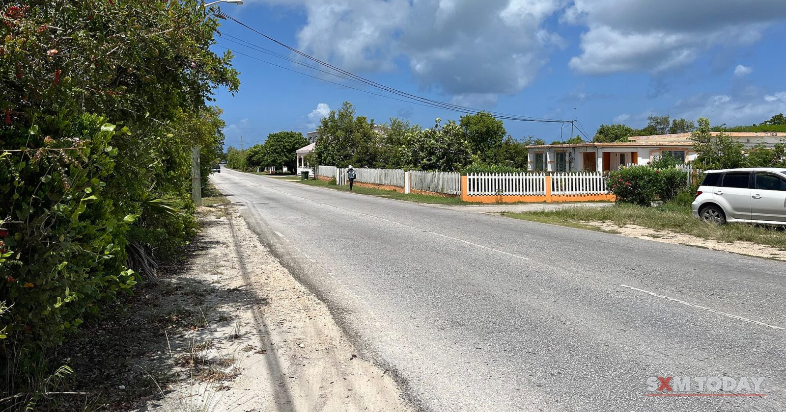 A straight paved road with a sandy verge runs past fenced single-storey houses and a parked white car under a blue sky with white clouds.