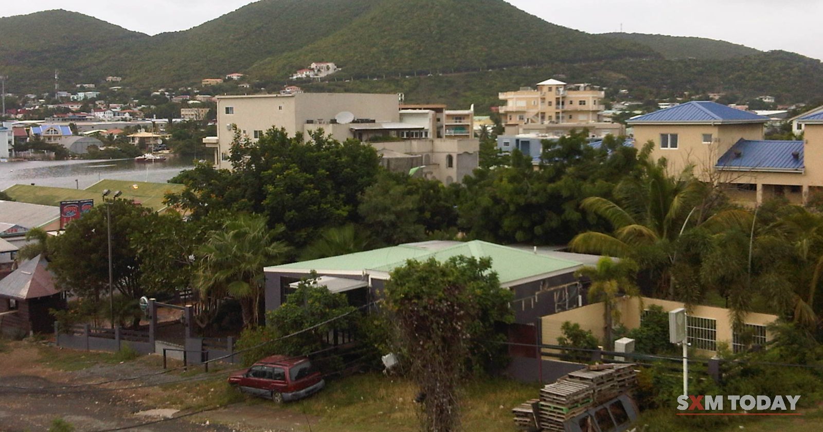 Low-rise houses and apartment blocks among trees below green hills under an overcast sky.