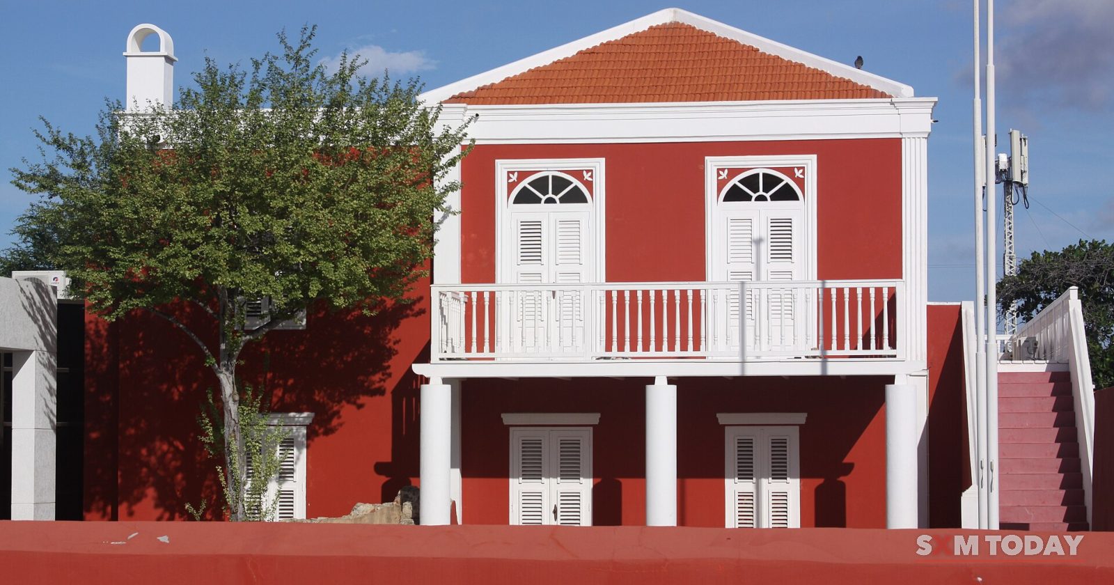 A red two-storey colonial building with white shutters, a white balcony and a red tile roof behind a red wall.