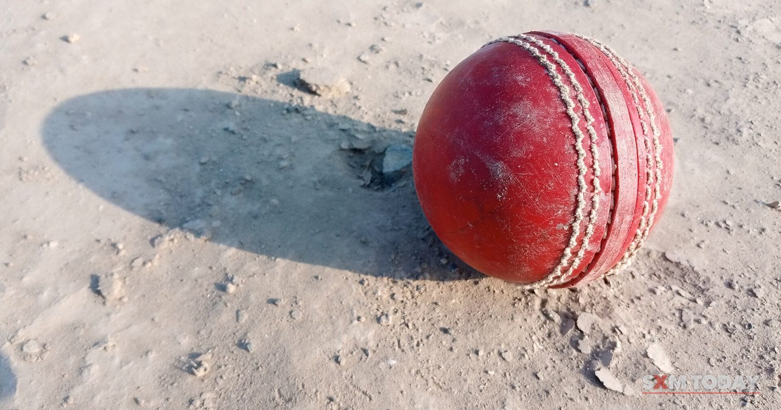 A red leather cricket ball with a raised seam sits on dry, sandy ground, casting a shadow.