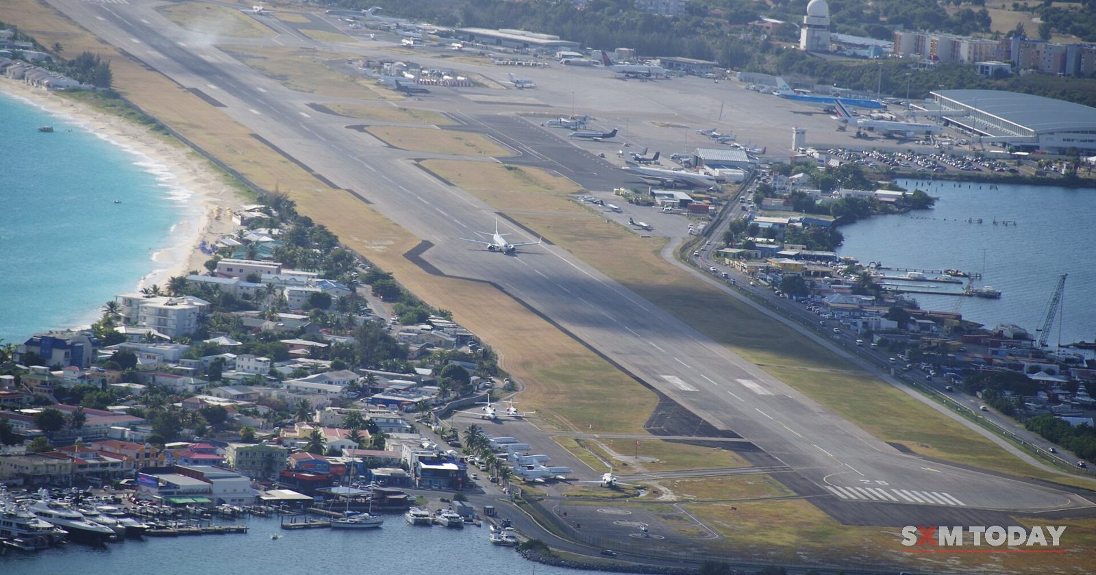 High view of an airport runway running along a white-sand beach, with the apron, parked planes and a lagoon beside it.