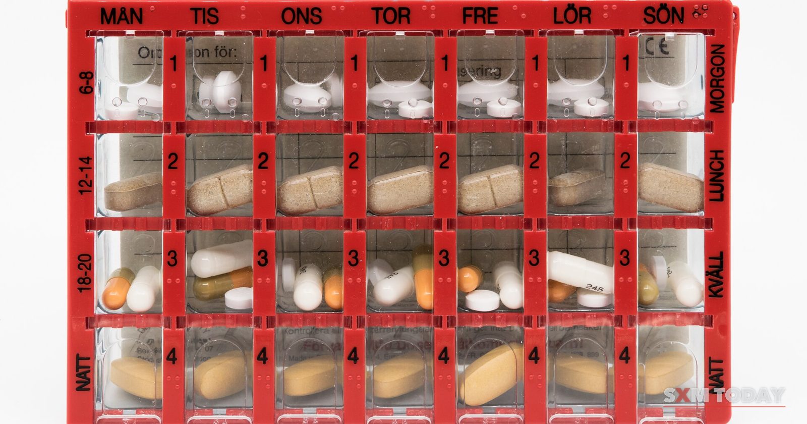A red weekly pill organiser with its compartments filled with tablets and capsules, on a white background.