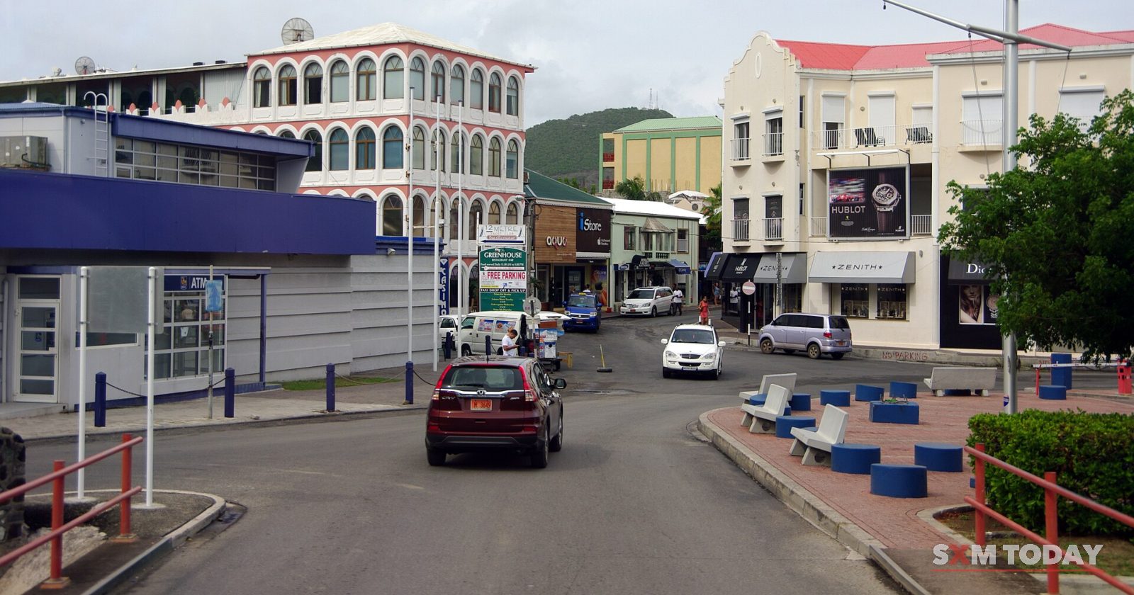 A street curves past arcaded buildings and a small park, with cars driving and parked under a cloudy sky.