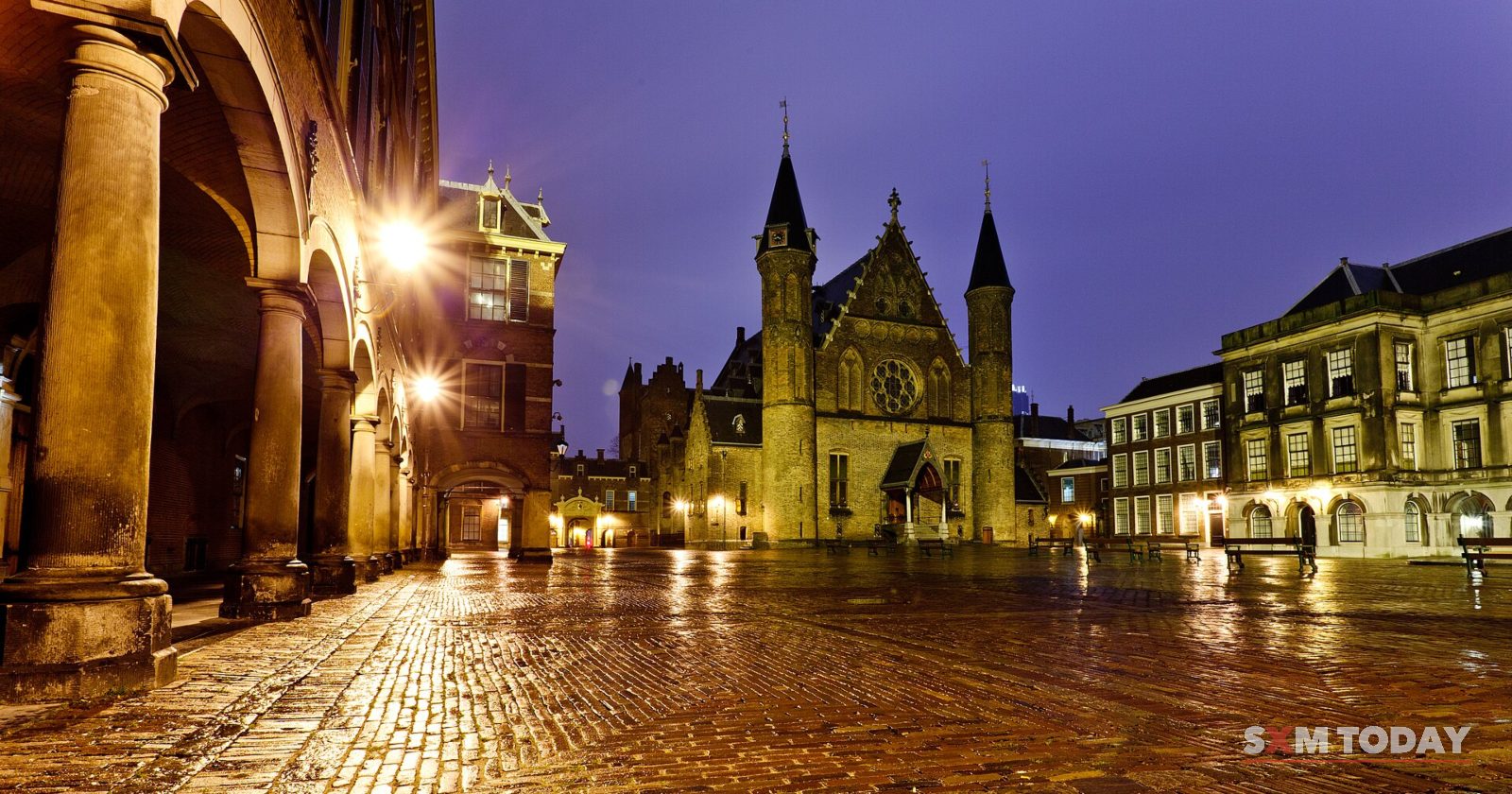 A Gothic hall with two spired towers and surrounding historic buildings lit at night, seen from a colonnade across a wet cobbled square.
