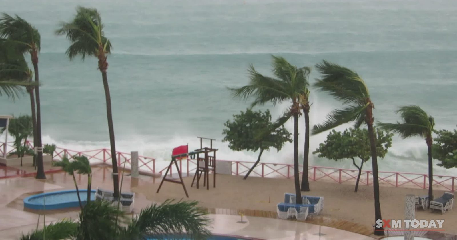 Palm trees bend in strong wind above a churning grey sea, with an empty pool deck and lifeguard tower below.