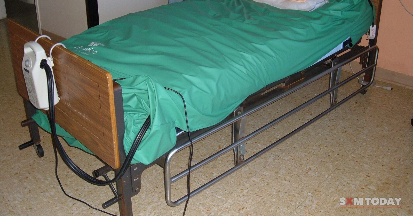 An adjustable care bed with a green pressure-relief mattress and a pump unit hooked on its footboard in a small room.