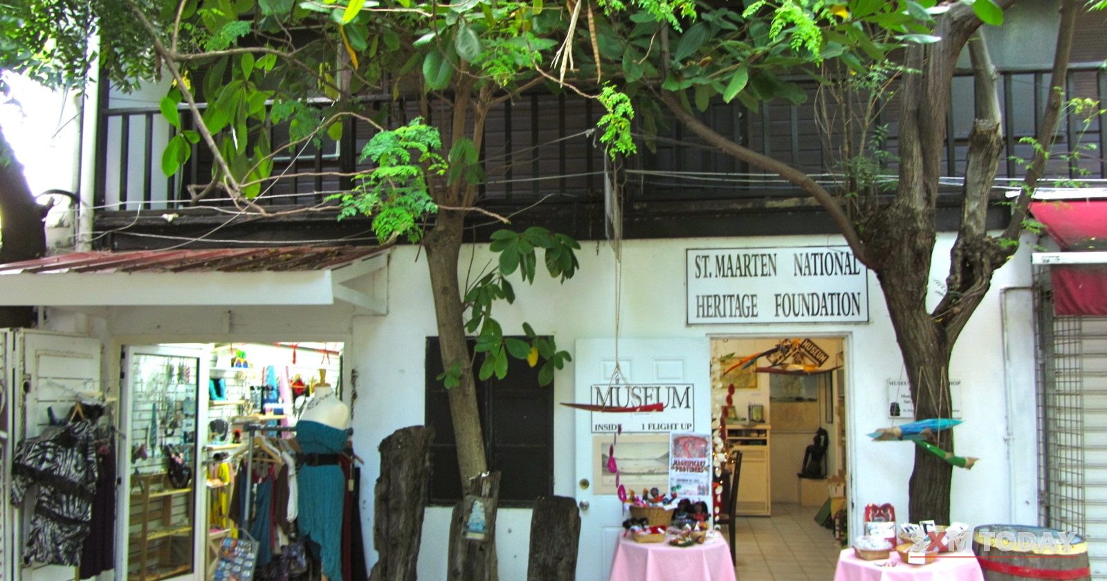 The entrance to a museum on a shaded street, with its sign above the door and a clothes rack and tables set out in front.