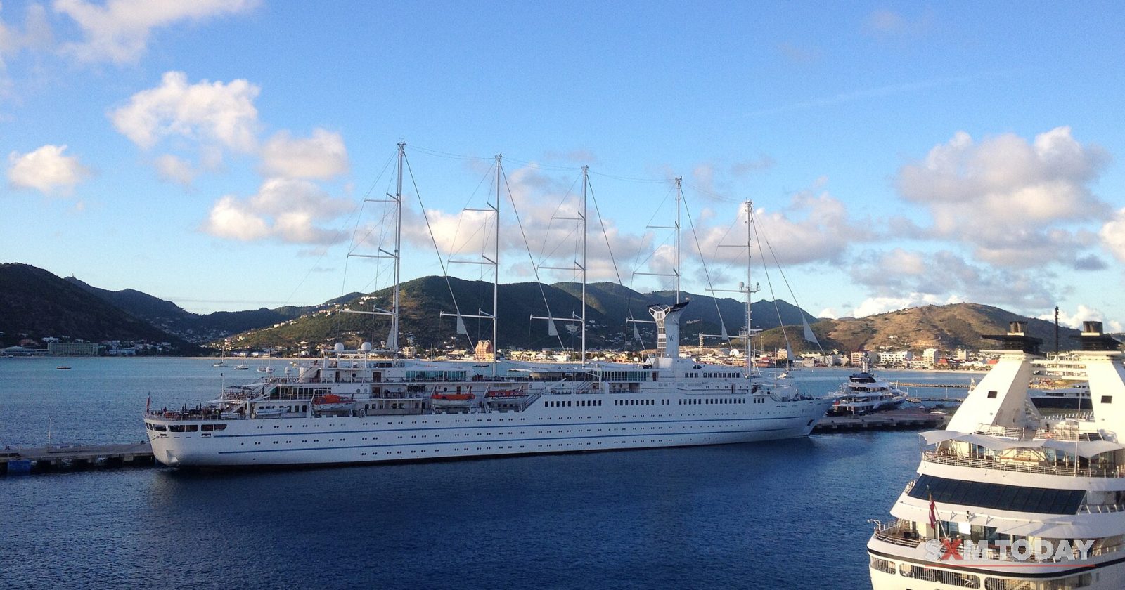 A four-masted cruise ship lies at a pier in deep blue water, with the bow of a second cruise ship in the foreground and green hills behind.