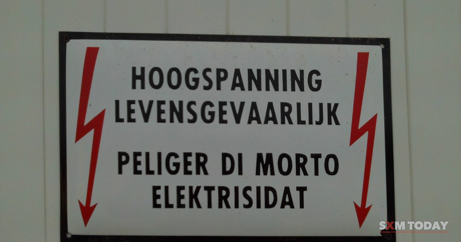 A white warning sign with red lightning arrows reads high voltage, danger of death, in Dutch and Papiamentu.