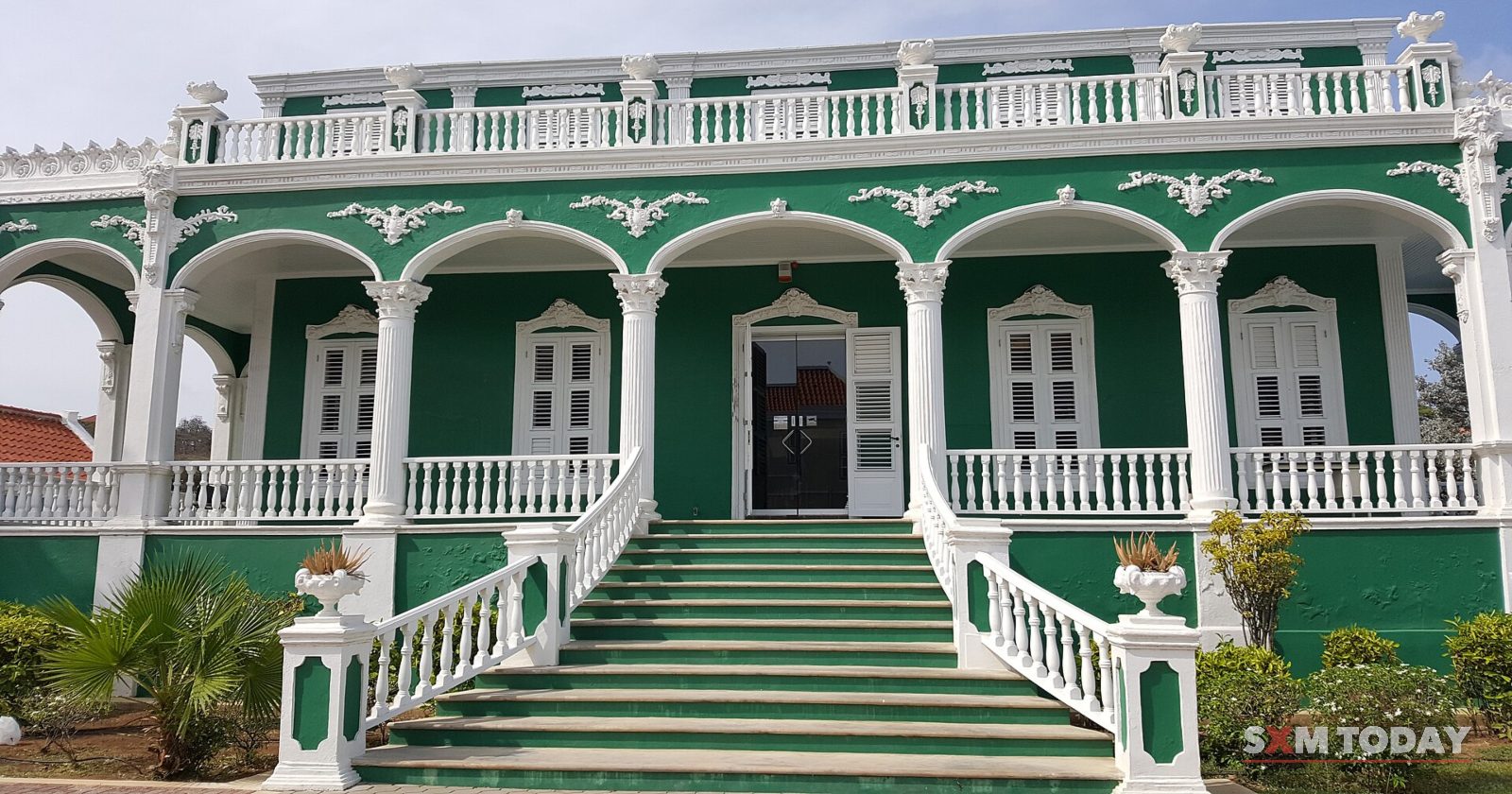 A green and white colonial building with arched verandas on two floors and a broad flight of steps up to its door.