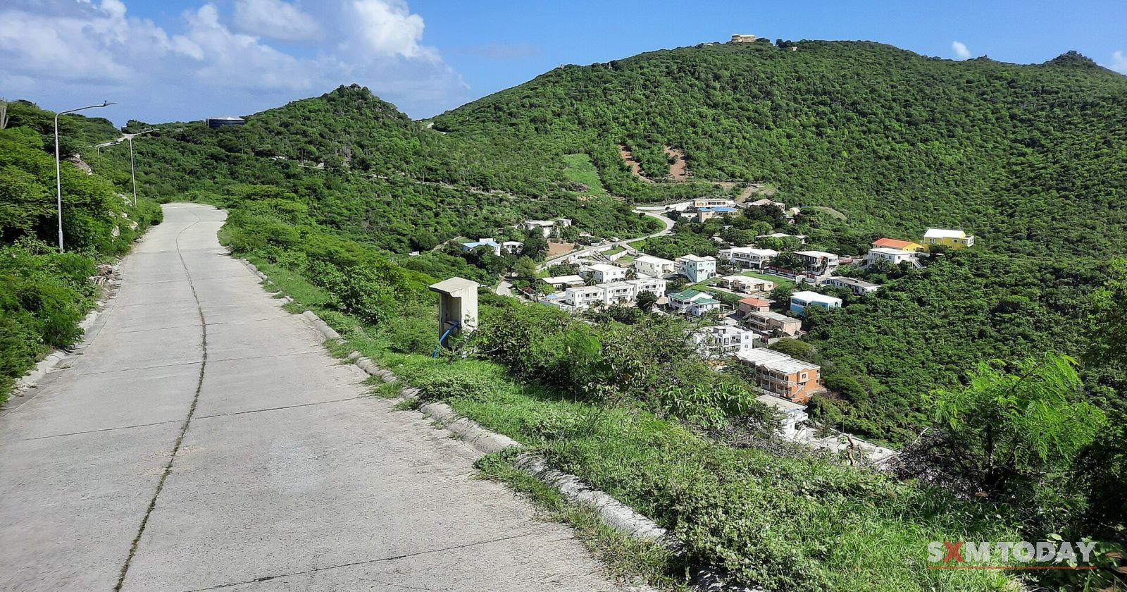 A narrow concrete road runs down a steep green hillside past scattered houses, with more hills behind.