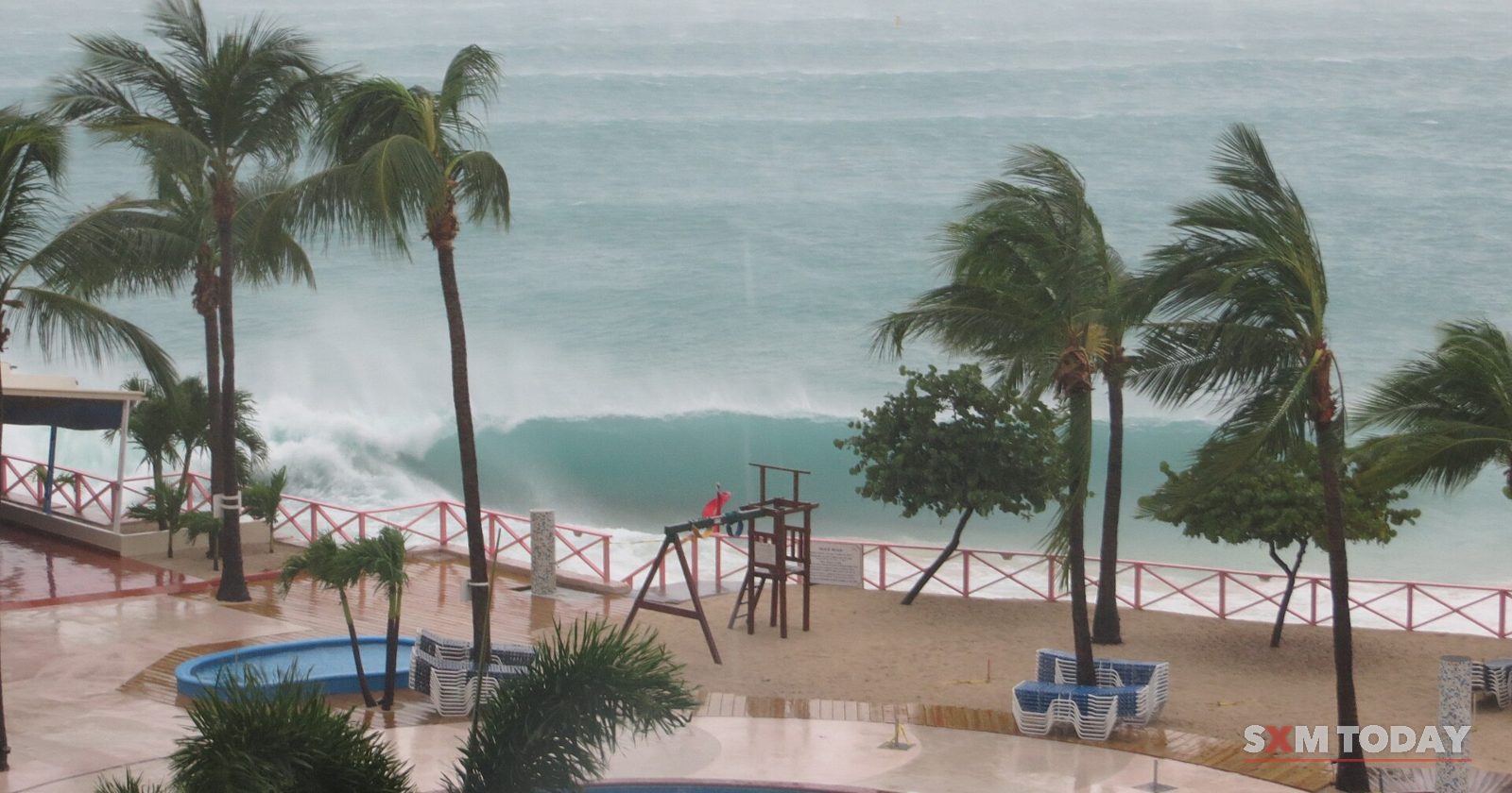 A large wave breaks behind wind-whipped palm trees above an empty beach, pool and lifeguard tower.