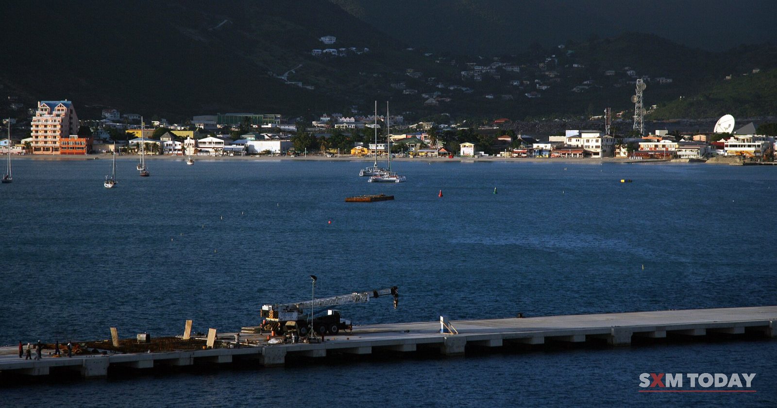 A pier with construction equipment in the foreground and a waterfront town below dark green hills across the bay.