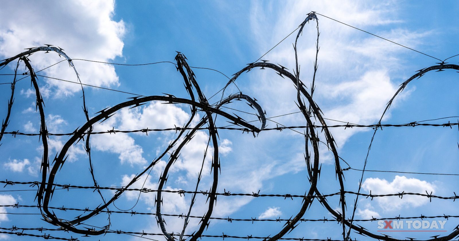Loops of razor wire and straight strands of barbed wire are silhouetted against a blue sky with white clouds.