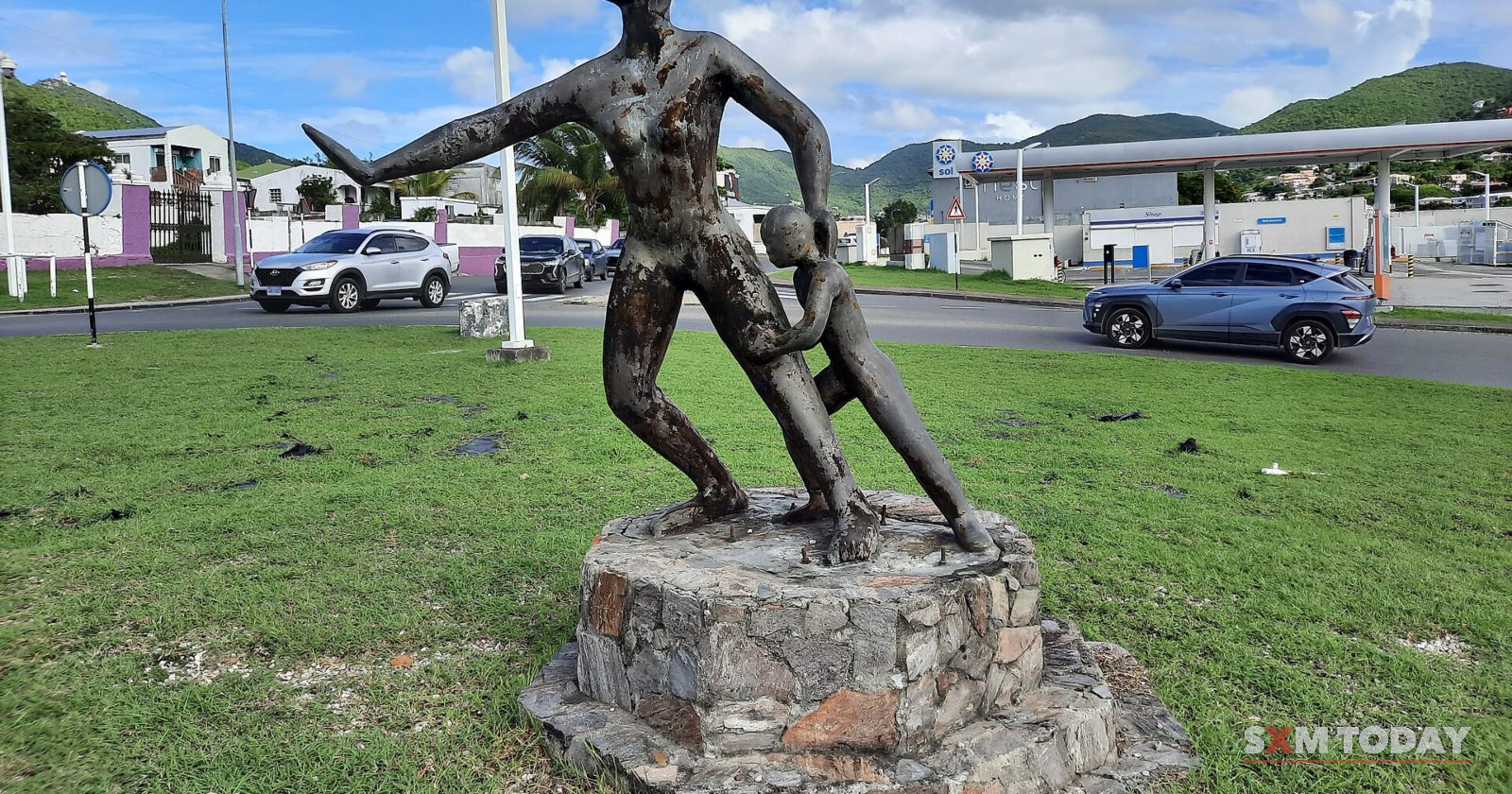 A dark metal sculpture of a figure shielding a child stands on a stone base on a grassy roundabout, with cars passing behind.