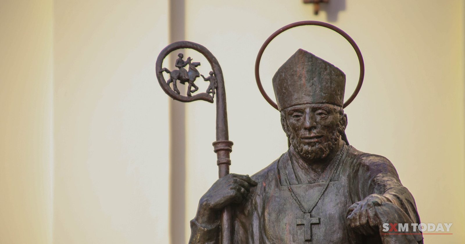 A bronze statue of a bishop with a halo, mitre and crozier against a pale wall.