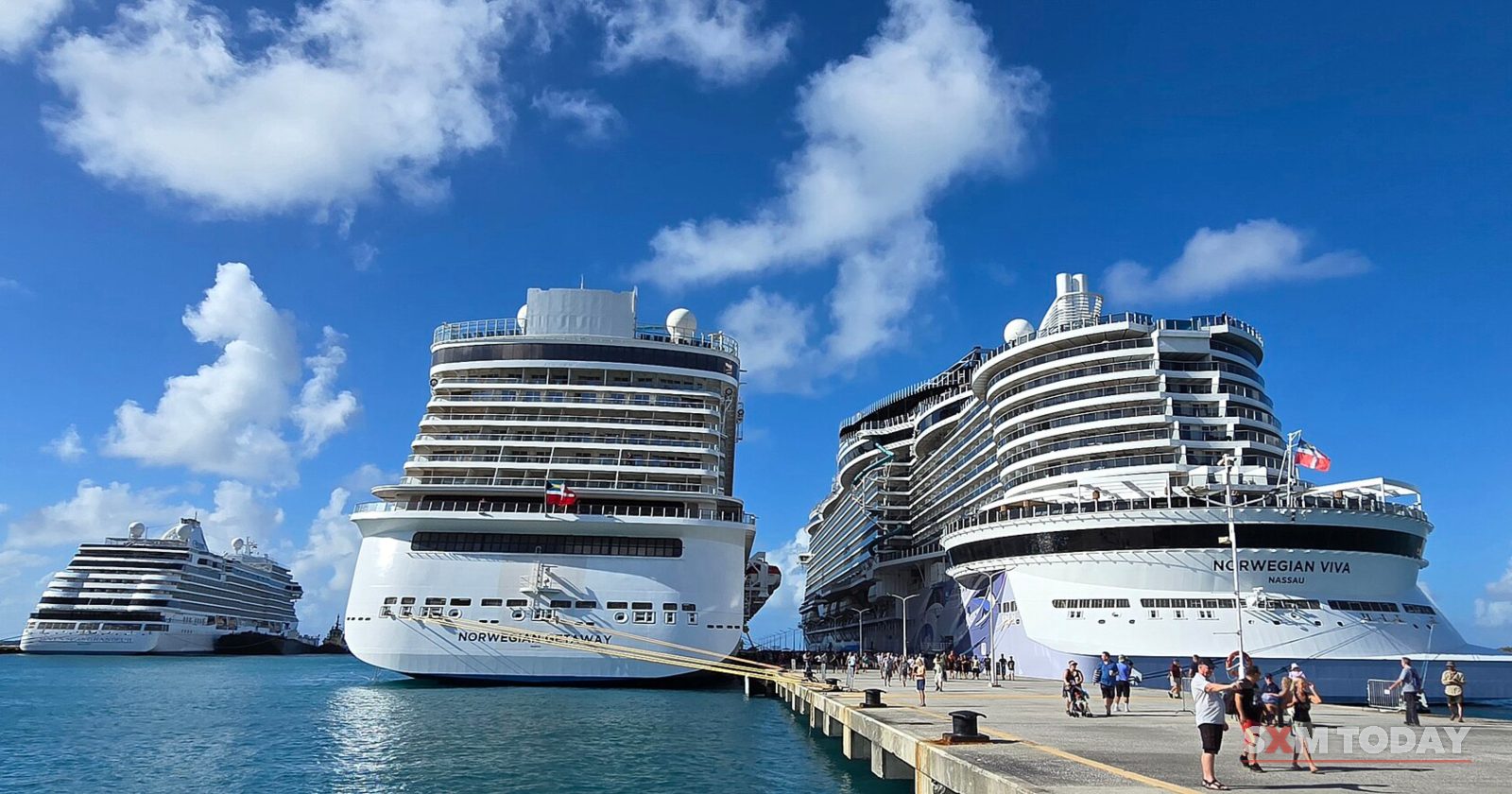 Three large cruise ships are berthed at a long pier under a blue sky with white clouds, with small figures walking along the quay.