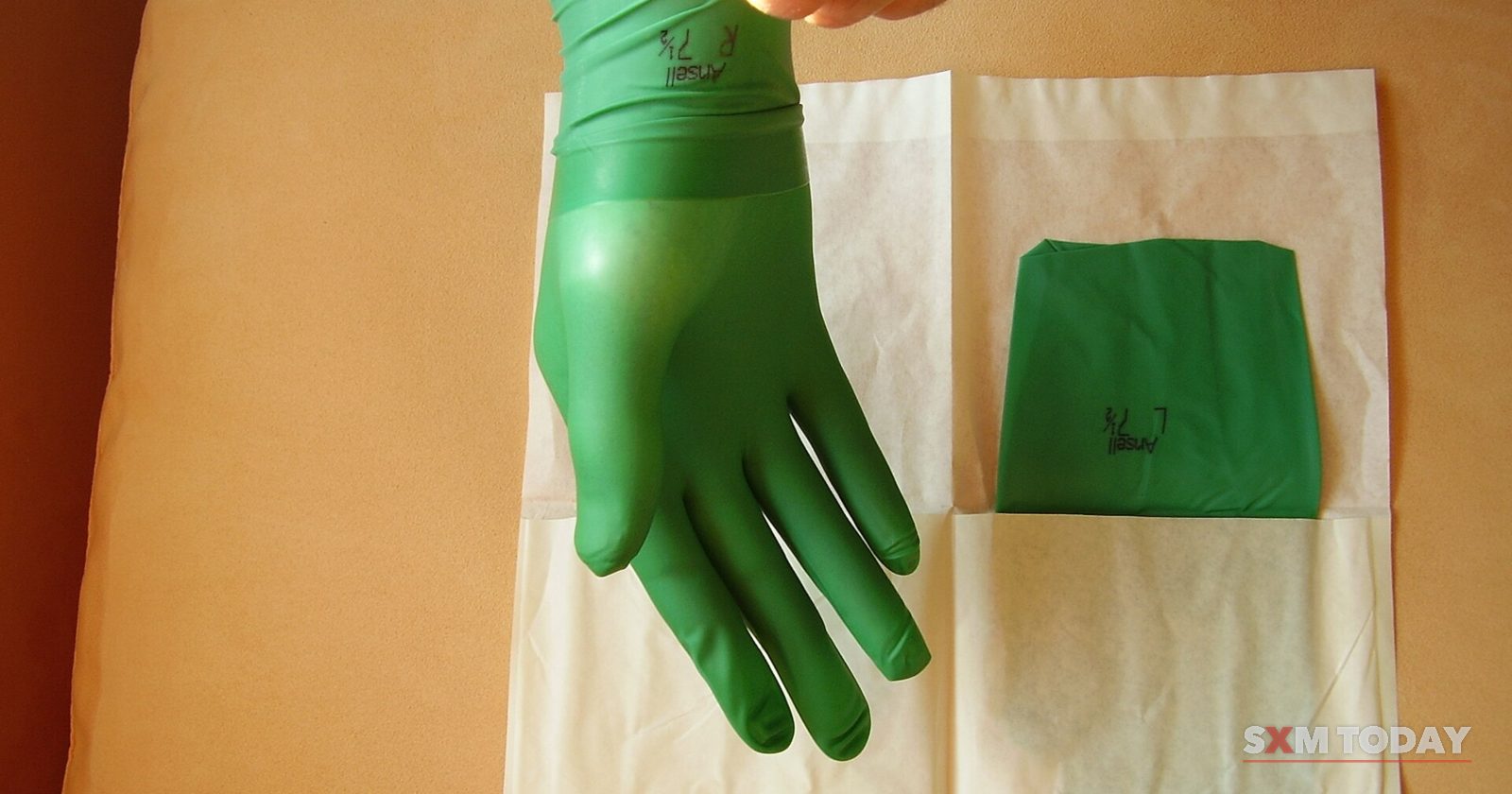 A hand wearing a green surgical glove beside an opened sterile wrapper holding the second glove.