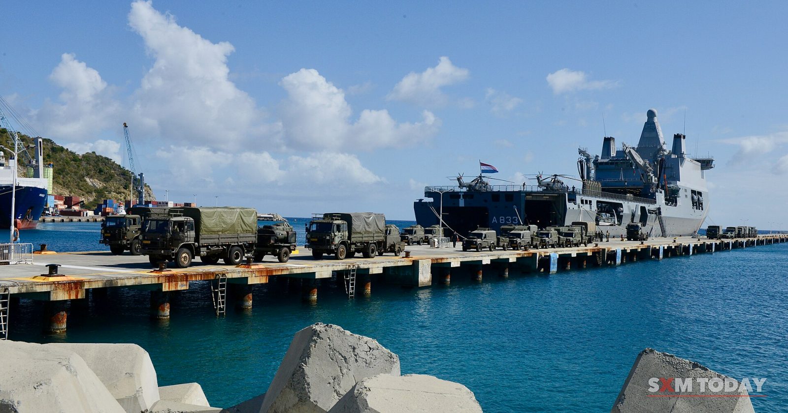 A grey naval ship moored at a long pier lined with military trucks, concrete breakwater blocks in the foreground.